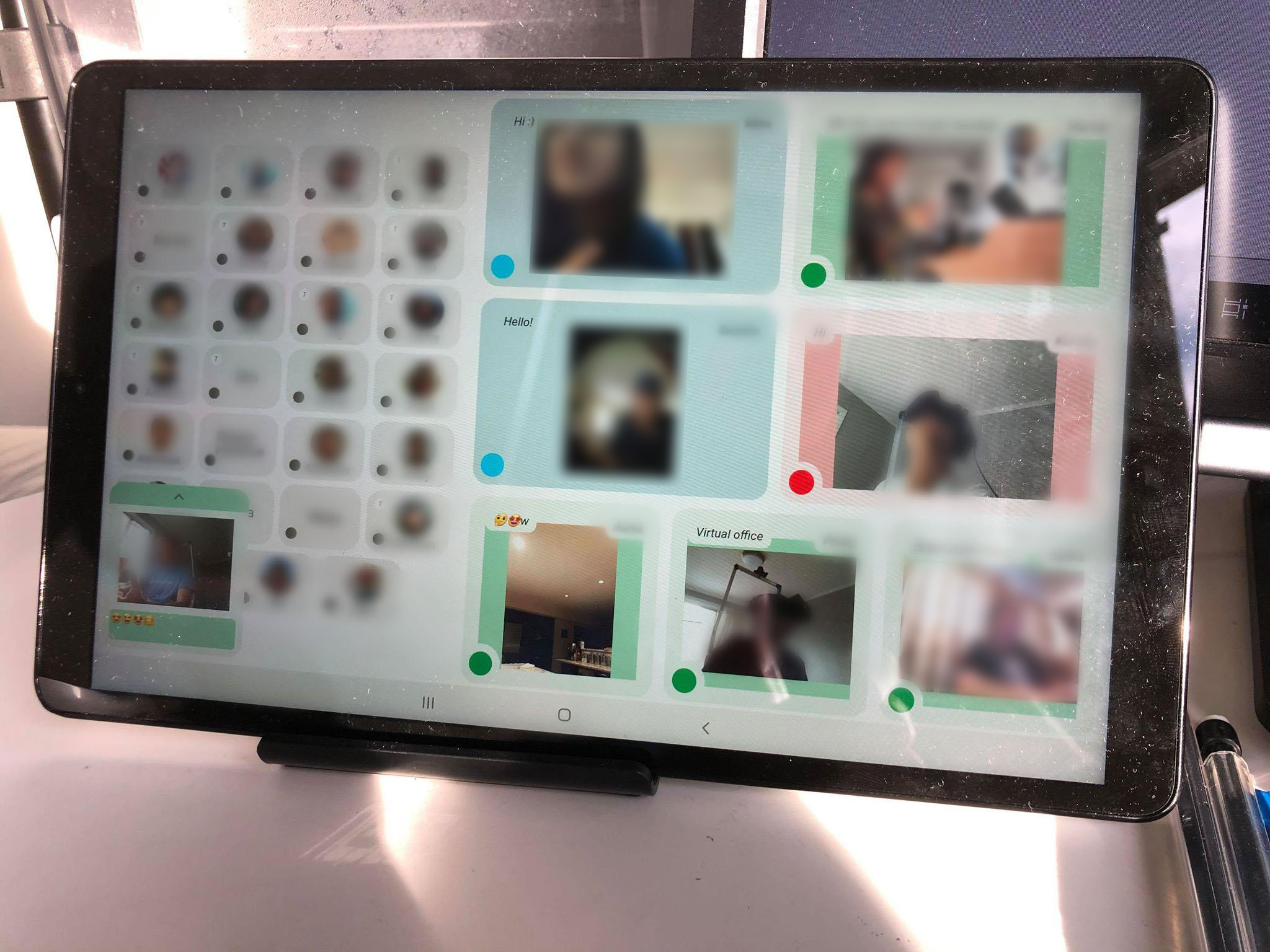 Photo of the tablet in use. Seven video feeds are displayed. Each is of a friend who is on camera and can also see you through the camera on the tablet. Each video has a colored border representing the status of the user. In the photo, four users are green, two are blue and one is red. There are also a number of gray regions with profile pictures depicting users who are offline. The photo is heavily blurred to protect the privacy of the users in the photo.